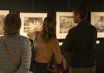 Tre personer ser på litografier av maritime motiver i en mørk utstillingssal. En kvinne peker på et av bildene.