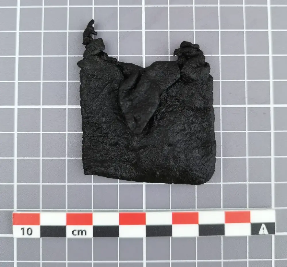 Liten firkantet lærveske fra middelalderen med snorøyer i toppen og en liten klaff foran, vist på rutet bakgrunn med målestokk.