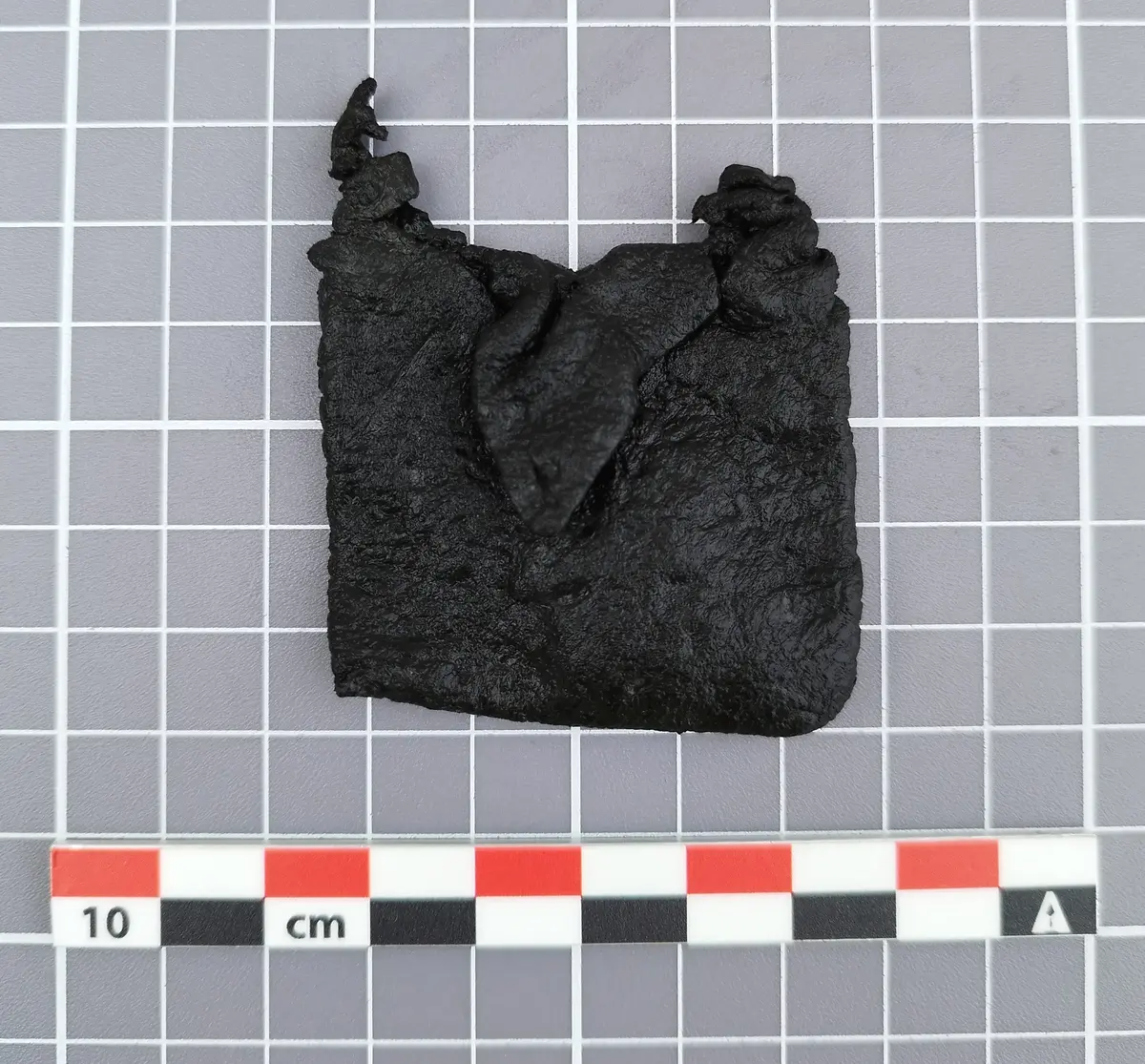 Liten firkantet lærveske fra middelalderen med snorøyer i toppen og en liten klaff foran, vist på rutet bakgrunn med målestokk.