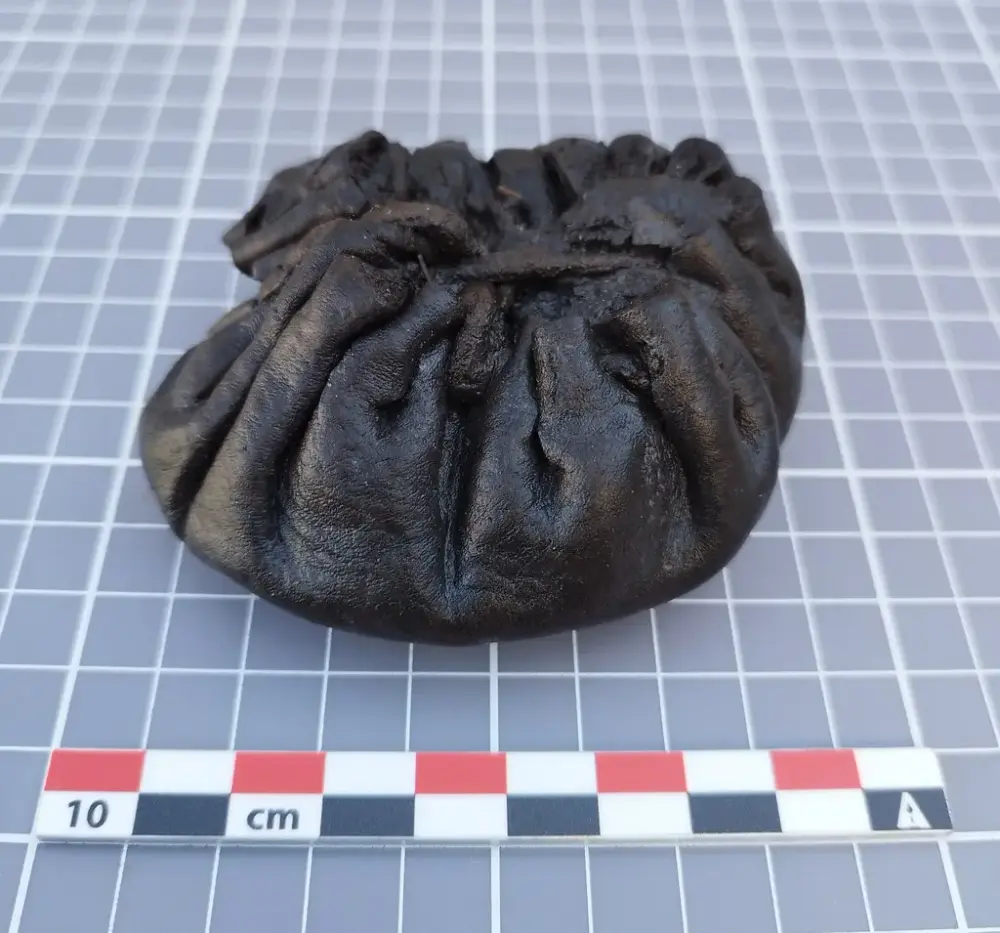 Liten lærpung fra middelalderen, rynket sammen i toppen med snøring, vist på rutet bakgrunn med målestokk.