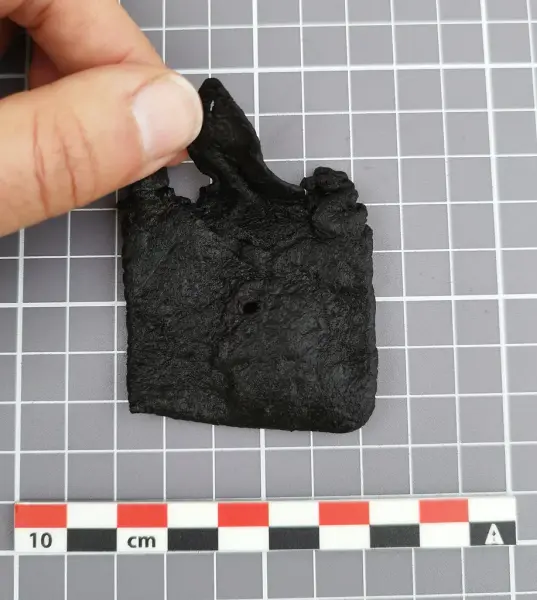 Liten lærveske med klaff og synlig hull midt på, holdt i hånden over rutet bakgrunn med målestokk.