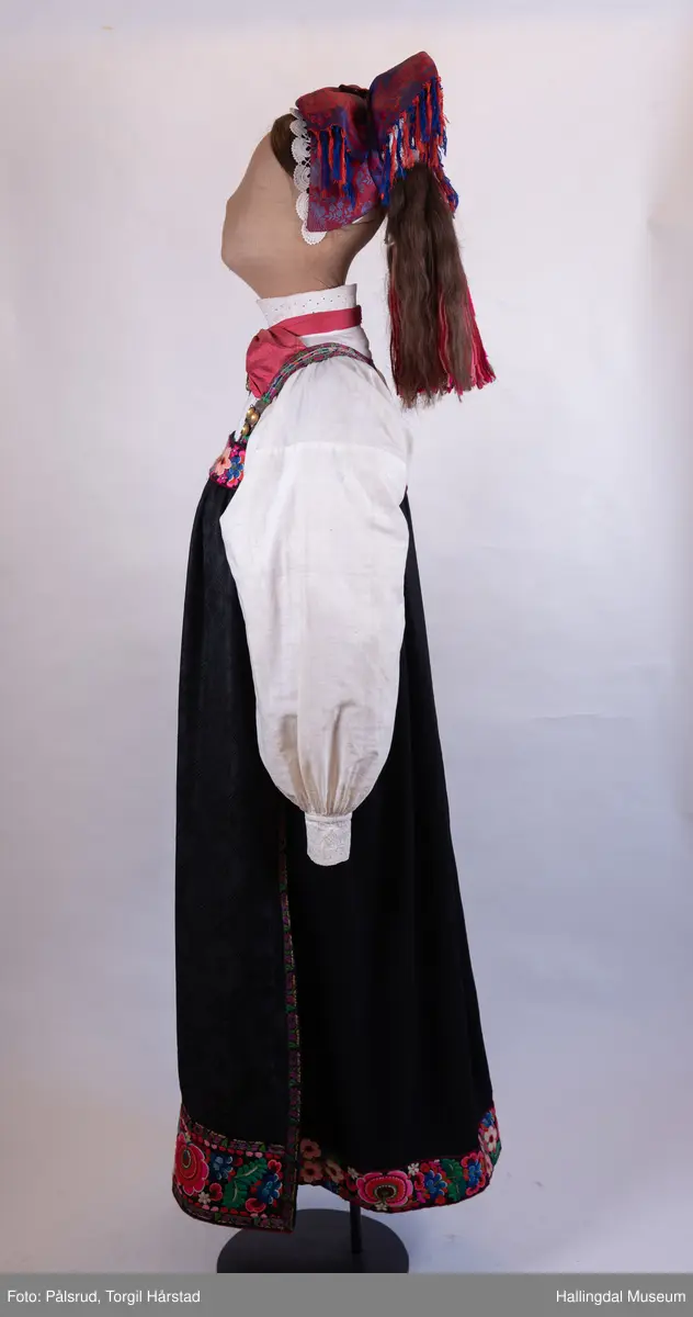 Kone frå Øvre Hallingdal, kyrkjekledd. Viser korleis gifte kvinner kunne gå kledd i Øvre Hallingdal. 

Truleg frå tidleg 1900-tal. Døs i svart ull, med brodert overliv i plattsaum og plattsaumbroderi nedst. forkle i svart ulldamask med plattsaumbroderi nedst. Døsa og forkle er truleg sauma av Birgit Anfinset (f. 1863). Sauma band kom nedanfrå dalen sist på 1800-talet, fyrst som forkleband. Banda var breiast rundt 1920, og denne stakken er truleg frå denne tida.

Hovudbunaden kallast hettepannelin eller lykkjeplagg (Ål), og er hovudbunaden til gifte. Her har silkeplagget (hettepannelinet) vorte stort og ferdigsydd. Storleiken på hettepannelinet har variert frå tid til anna.

Kona står utstilt i den faste bunadsutstillinga på Hol Bygdemuseum. 
