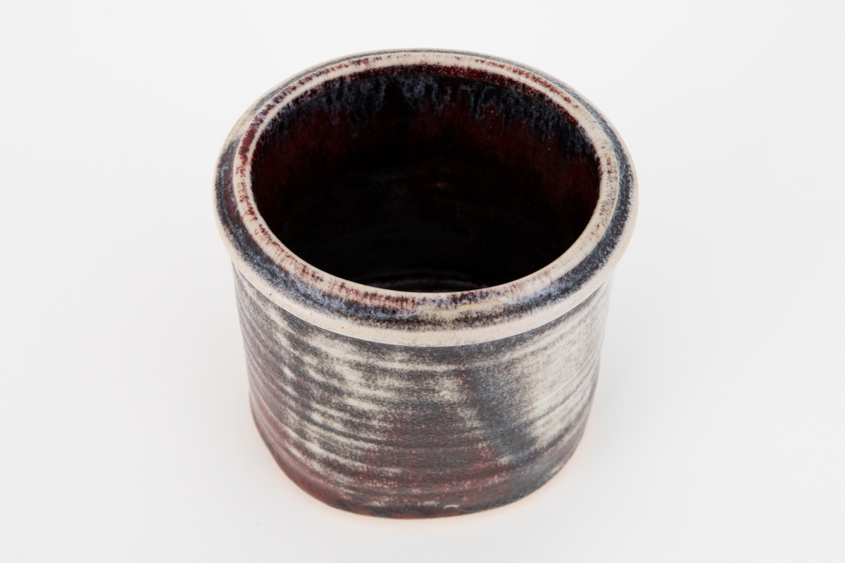 Krukke med oval form i glasert steingods. Godset skinner delvis gjennom den halvblanke glasuren, som er i brune og gråhvite toner.  Krukken har rette vegger.