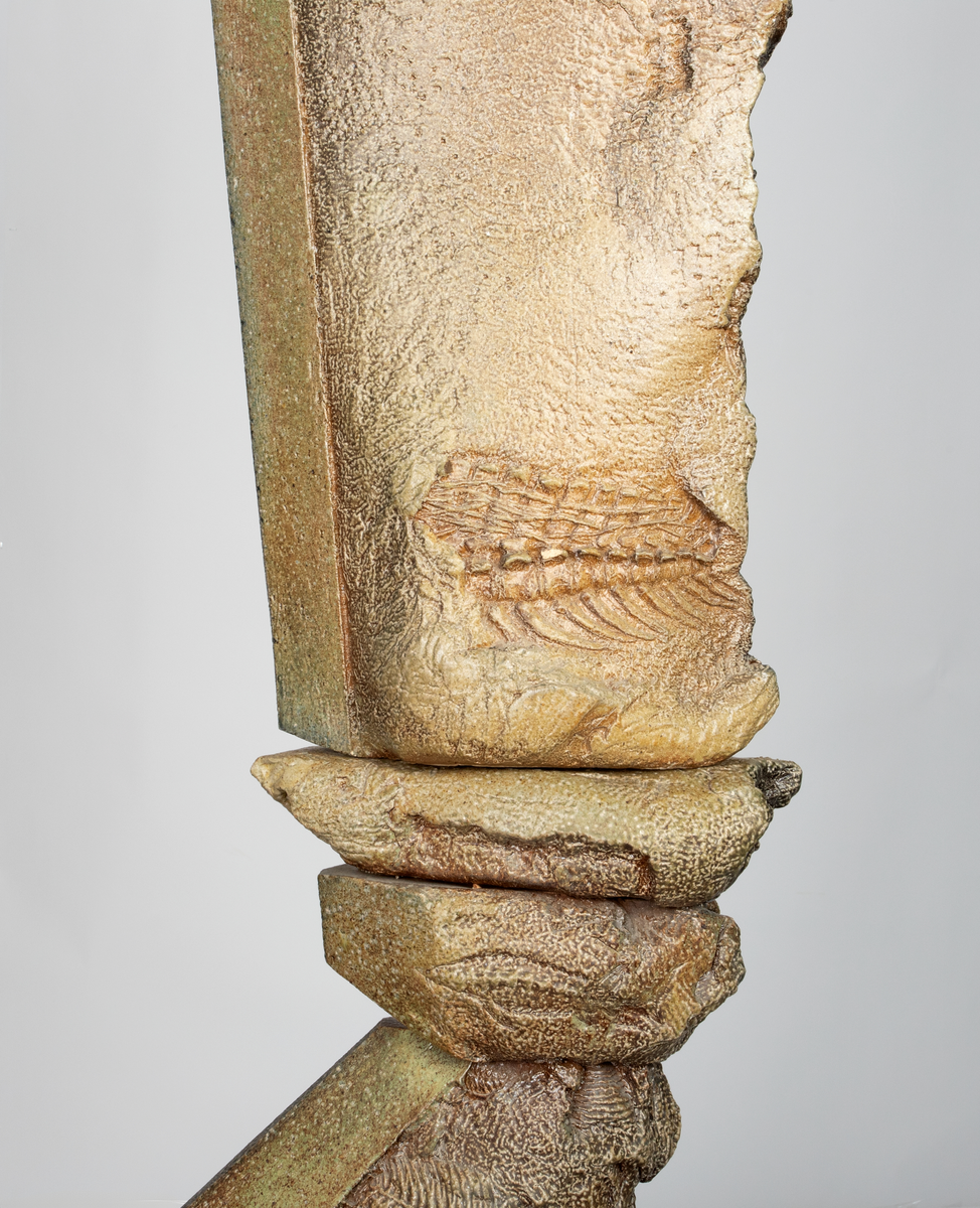 Keramisk skulptur på sort sokkel med sand. Skulpturen består av organiske former som stiger opp av sanda. I formene er det avtrykk som forestiller fossiler, avtrykk av bein og abstrakte former. Skulpturen er glasert beige og brun.