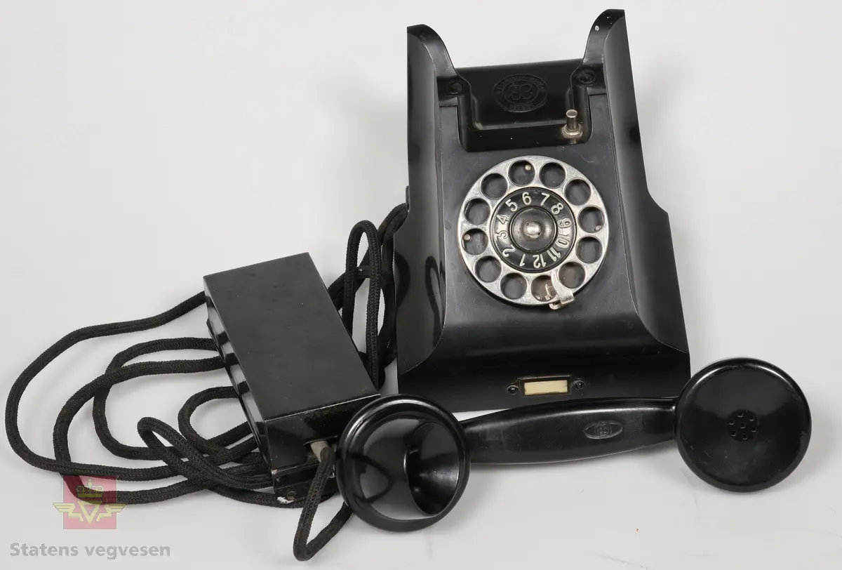 Svart telefonapparat med sølv/grått hjul på framsiden til å velge nummer. Markert med Logo EB (elektrisk Bureau). 