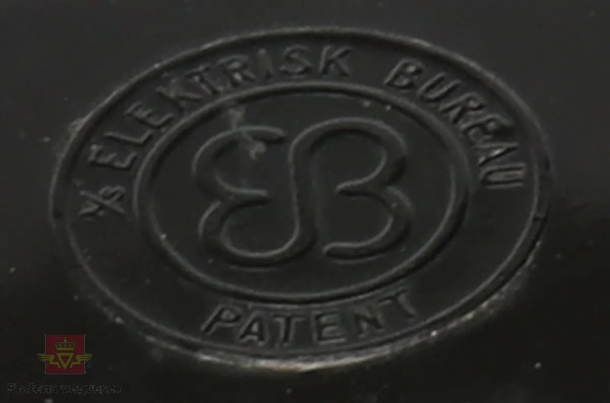 Svart telefonapparat med sølv/grått hjul på framsiden til å velge nummer. Markert med Logo EB (elektrisk Bureau). 