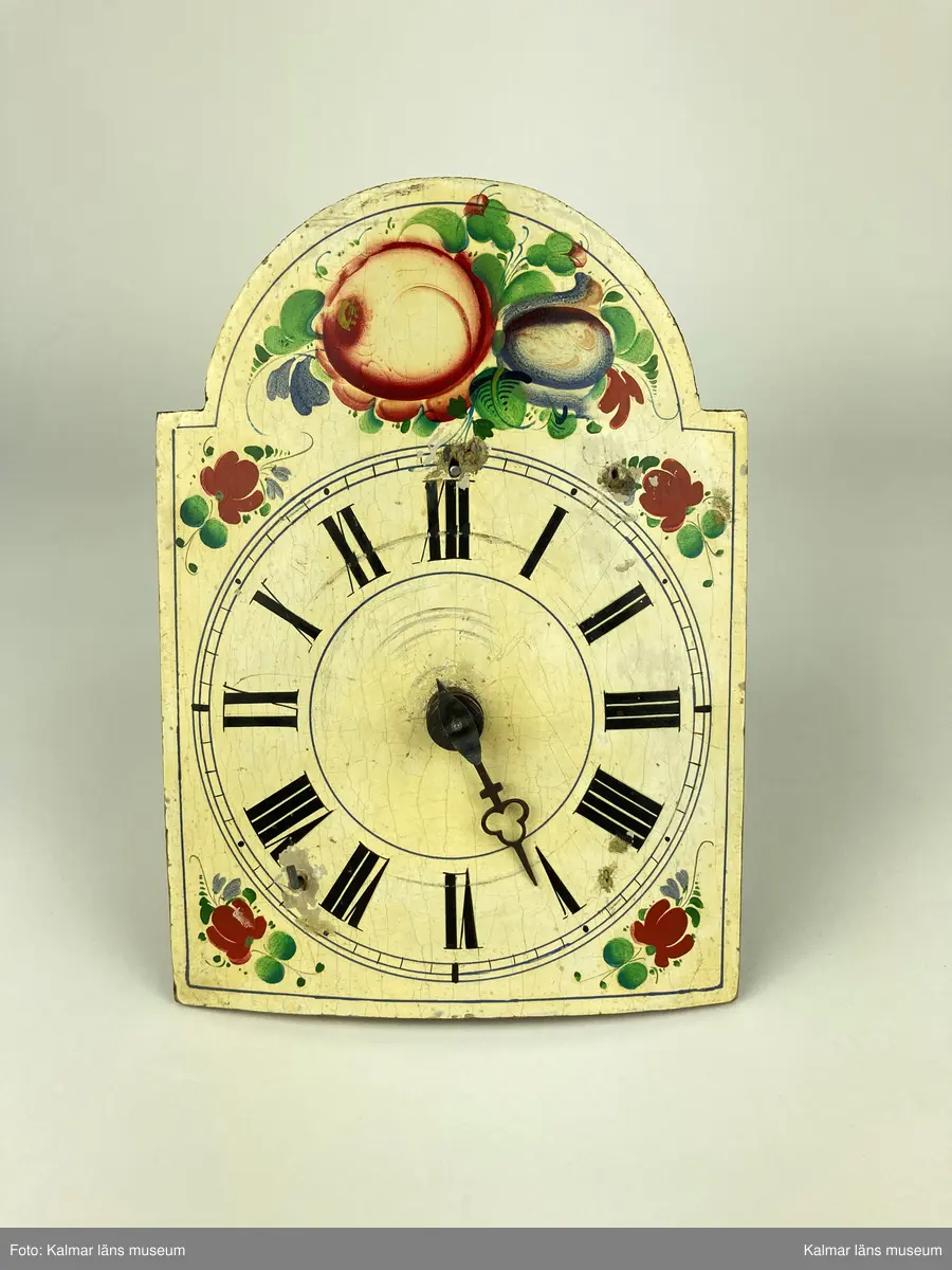 KLM 10433. Väggur. Urtavla av trä målad i vitt med romerska siffror i svart samt blommor och blad i grönt, rött och blått. Urverk av mässing och stål innefattad i ett träfodral med öppningsbara luckor på sidorna. En visare är lös. Lod, lodlinor och pendel saknas.