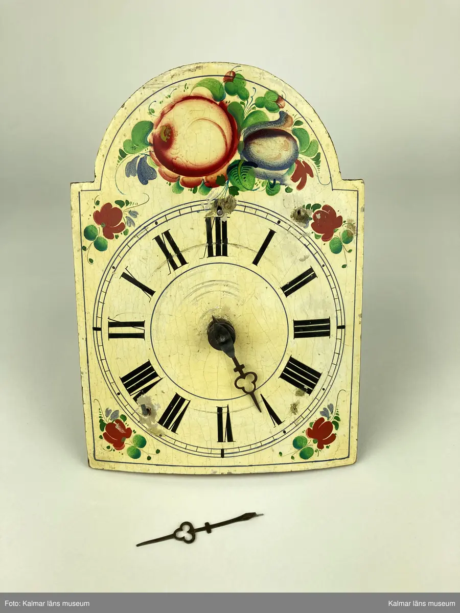KLM 10433. Väggur. Urtavla av trä målad i vitt med romerska siffror i svart samt blommor och blad i grönt, rött och blått. Urverk av mässing och stål innefattad i ett träfodral med öppningsbara luckor på sidorna. En visare är lös. Lod, lodlinor och pendel saknas.