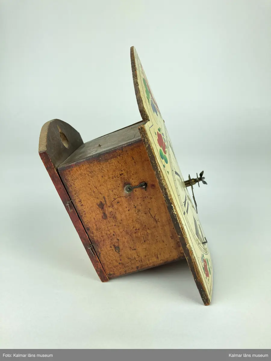 KLM 10433. Väggur. Urtavla av trä målad i vitt med romerska siffror i svart samt blommor och blad i grönt, rött och blått. Urverk av mässing och stål innefattad i ett träfodral med öppningsbara luckor på sidorna. En visare är lös. Lod, lodlinor och pendel saknas.