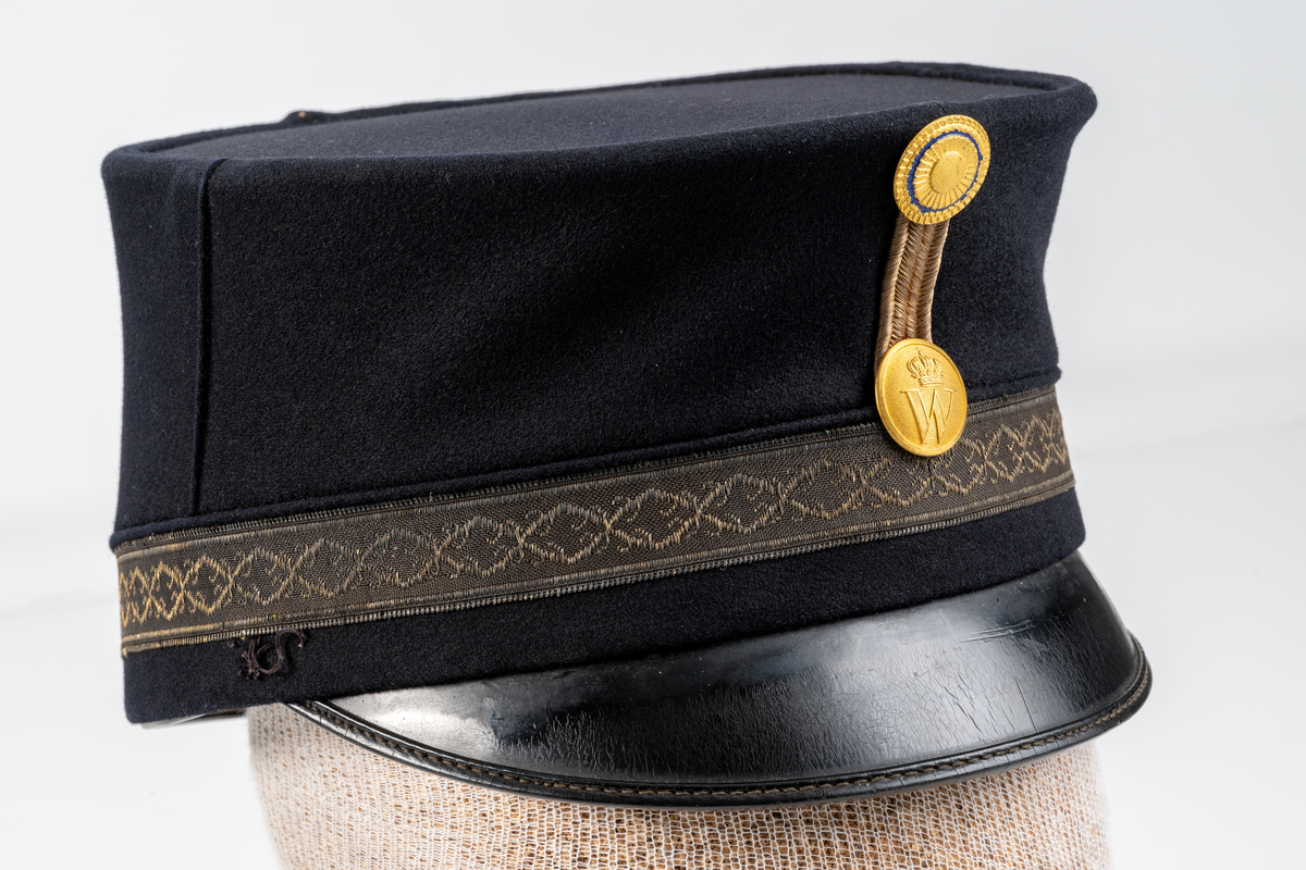 Skärmmössa (modell högmössa) i mörkblått kläde. Raka sidor med platt kulle. Mönstervävt mössband. På mössans framsida, agraff med rund kokard i mässing och undertill en mössknapp präglad med Växjö stadsvapen (år 1872-1926). Svart lackad skärm. Mössan saknar hakband med två knappar. Svettrem i brunt läder. Fodrad med svart tyg.