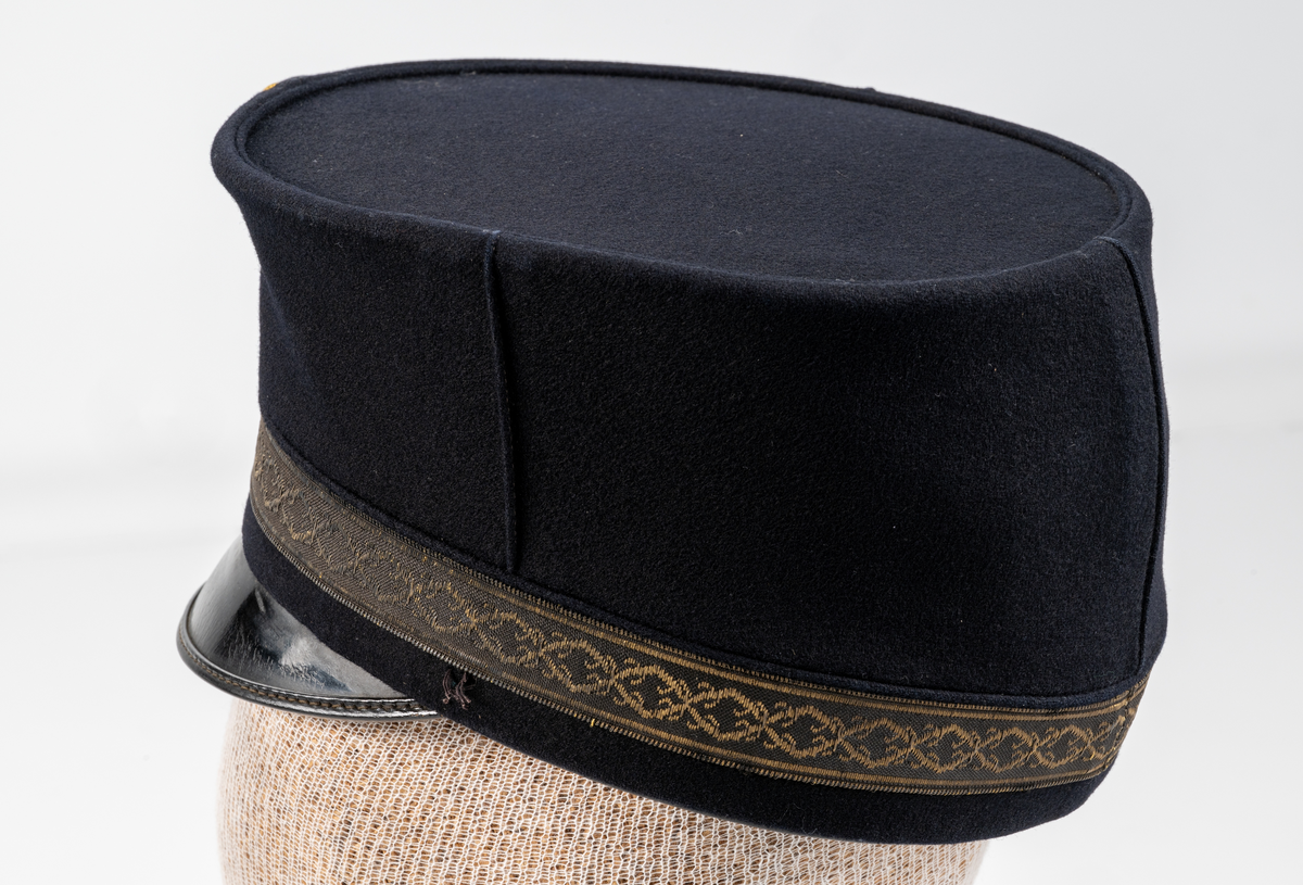 Skärmmössa (modell högmössa) i mörkblått kläde. Raka sidor med platt kulle. Mönstervävt mössband. På mössans framsida, agraff med rund kokard i mässing och undertill en mössknapp präglad med Växjö stadsvapen (år 1872-1926). Svart lackad skärm. Mössan saknar hakband med två knappar. Svettrem i brunt läder. Fodrad med svart tyg.