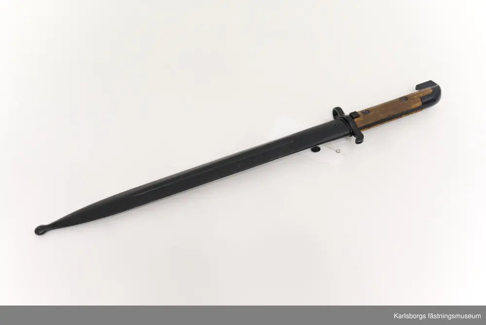 Bajonett m/1914, för karbin m/1894-1914. Bajonett är ett blankvapen som fästs vid mynningen på musköter, gevär eller karbiner och som används i närstrid. Karbin m/1894-1914 saknade ursprungligen bajonett, men försågs år 1914 med sådan. Märkt EJ AB (Eskilstuna Jernmanufaktur AB). Bajonetten kommer med bajonettbalja. Handtag av metall och trä.
