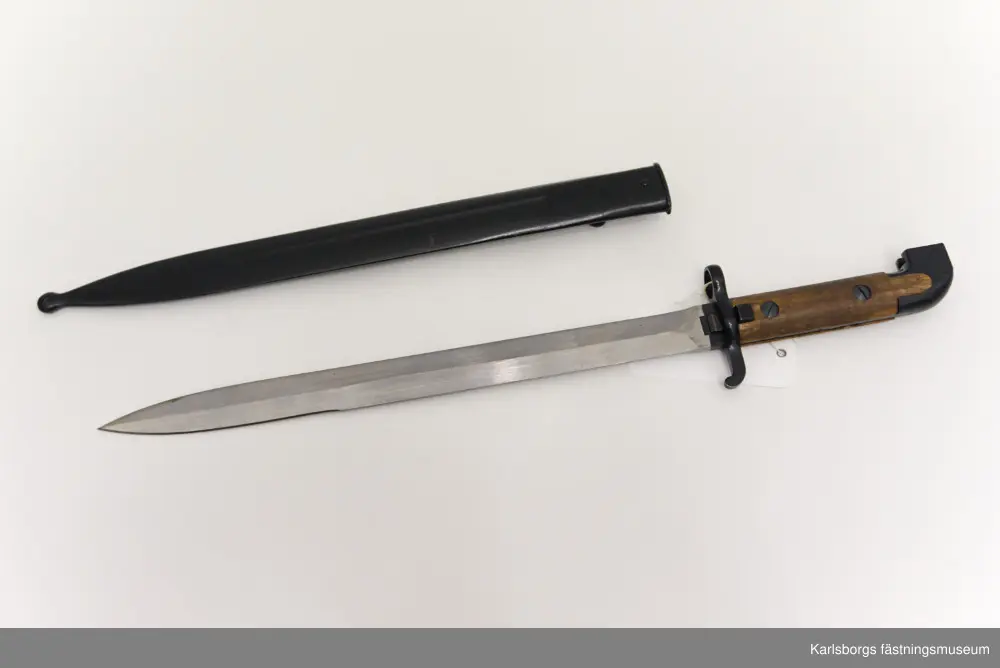 Bajonett m/1914, för karbin m/1894-1914. Bajonett är ett blankvapen som fästs vid mynningen på musköter, gevär eller karbiner och som används i närstrid. Karbin m/1894-1914 saknade ursprungligen bajonett, men försågs år 1914 med sådan. Märkt EJ AB (Eskilstuna Jernmanufaktur AB). Bajonetten kommer med bajonettbalja. Handtag av metall och trä.