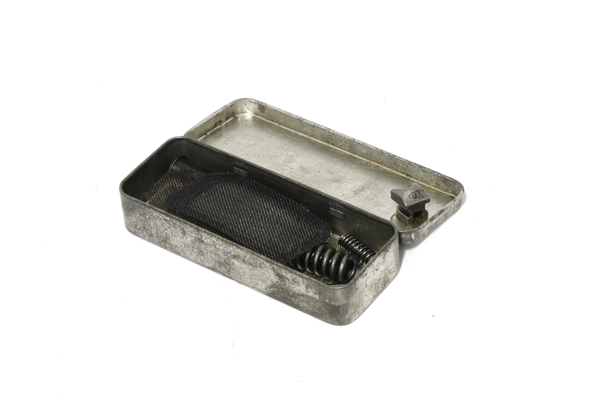 Rektangulær boks med hengslet lokk, inni ligger 6 løse deler:
2 fjærer, 1 sylinder, 2 nettinger, 1 liten metalldel.
Tilhører et sett med utstyr for rengjøring og istandsetting av våpen.