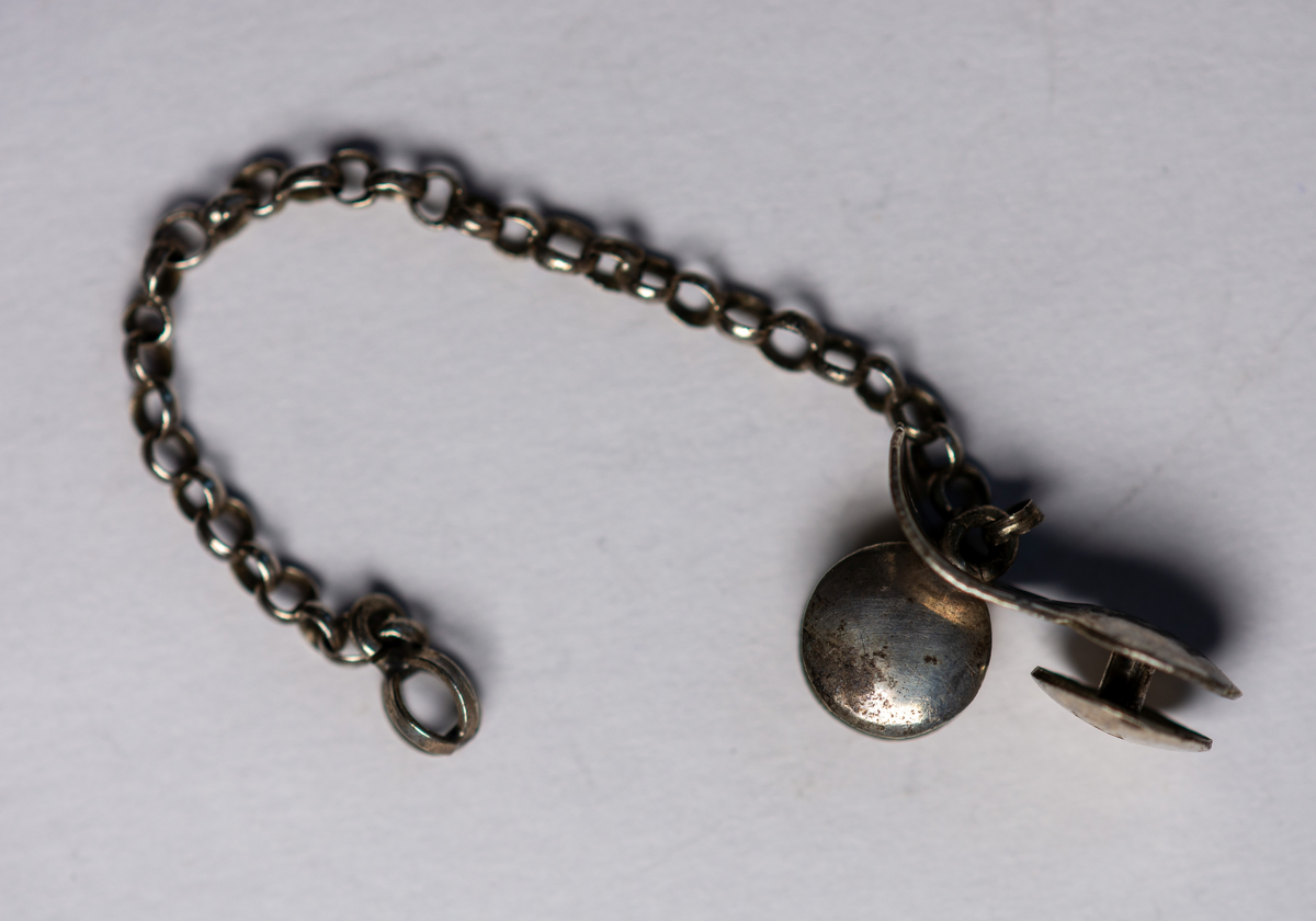 Kedja, fäst vid en stolpknapp. En stolpknapp rörlig längs kedjan. Silver .Längd 7,5 cm. (kedja)Diameter 0,8 cm (knappar)Stämplar: se bildZ5=1878