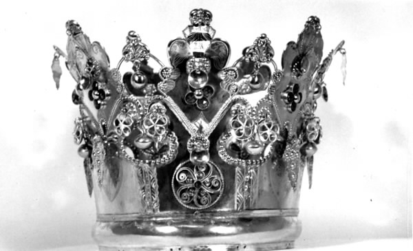 Brudkrona av silverplåt. Oval rak sida av 7 hoplödda plåtar som upptill svänger ut och avslutas i treflikade krön. Buckelornamentik samt svisselerade mönster. Fyrpassformade infattningar av röda och gröna glasstenar på varje flik. Ytterligare dekorerad med påsatta löst hängande förgyllda löv samt halvsfäriska hängen. Kronan avslutas nertill med en påsatt vulst. Stämplar: 13 1/3 (lödighet), LMS (=LM Salvesen), (Ålesunds stadsvapen), 1854.Mått upptill: längd 23 cm, bredd 23 cm.nertill: längd 17 cm, bredd 16 cm, höjd 17 cm.