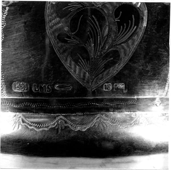 Brudkrona av silverplåt. Oval rak sida av 7 hoplödda plåtar som upptill svänger ut och avslutas i treflikade krön. Buckelornamentik samt svisselerade mönster. Fyrpassformade infattningar av röda och gröna glasstenar på varje flik. Ytterligare dekorerad med påsatta löst hängande förgyllda löv samt halvsfäriska hängen. Kronan avslutas nertill med en påsatt vulst. Stämplar: 13 1/3 (lödighet), LMS (=LM Salvesen), (Ålesunds stadsvapen), 1854.Mått upptill: längd 23 cm, bredd 23 cm.nertill: längd 17 cm, bredd 16 cm, höjd 17 cm.