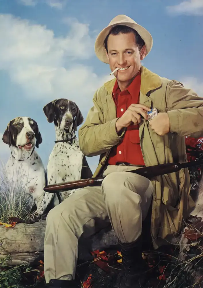 En smilende mann med sigarett i munnviken, gevær i fanget og to fuglehunder som ser på