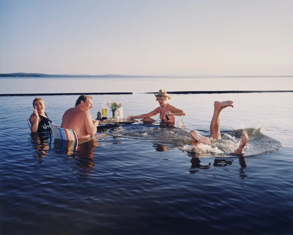 Fire personer sitter rundet et bord som er nedsenket i vann, en av dem er i ferd med å velte og bare beina stikker opp av vannet