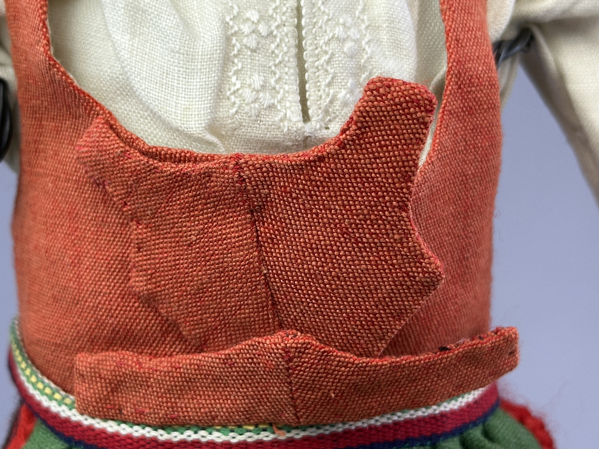 Docka klädd i kvinnlig sockendräkt från Norra Ny i Värmland. Kroppen är sydd i linnetyg och stoppad med textilt material. Huvud och händer är av lergods. Kläder sydda av handvävda tyger förutom skjorta, underkjol och halskläde, som är maskinvävt. Halskläde är broderat med tambursöm, skjortan med plattsöm och myrgång samt har stripade veck runt halskrage och manschetter. Även kjolen har lagda veck runtom och är fastsytt med livstycket. Förklädet, med 3 lagda veck på var sida, är kantad runtom med en tre-fläta gjord av rött ullgarn, vilket också kjolen har i nederkanten. Ullstrumporna är handstickade i slätstickning med en kant upptill i 2-2 resår och broderad med förstygn av rött ullgarn. Huvudet är prytt med hår av lintågor, som delvis är flätat till hårflätor samt ett målat hårnäver. Dockans fötter bär svarta skinnskor.