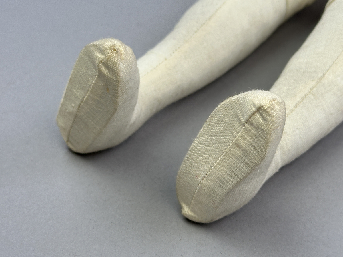 Docka klädd i kvinnlig sockendräkt från Norra Ny i Värmland. Kroppen är sydd i linnetyg och stoppad med textilt material. Huvud och händer är av lergods. Kläder sydda av handvävda tyger förutom skjorta, underkjol och halskläde, som är maskinvävt. Halskläde är broderat med tambursöm, skjortan med plattsöm och myrgång samt har stripade veck runt halskrage och manschetter. Även kjolen har lagda veck runtom och är fastsytt med livstycket. Förklädet, med 3 lagda veck på var sida, är kantad runtom med en tre-fläta gjord av rött ullgarn, vilket också kjolen har i nederkanten. Ullstrumporna är handstickade i slätstickning med en kant upptill i 2-2 resår och broderad med förstygn av rött ullgarn. Huvudet är prytt med hår av lintågor, som delvis är flätat till hårflätor samt ett målat hårnäver. Dockans fötter bär svarta skinnskor.