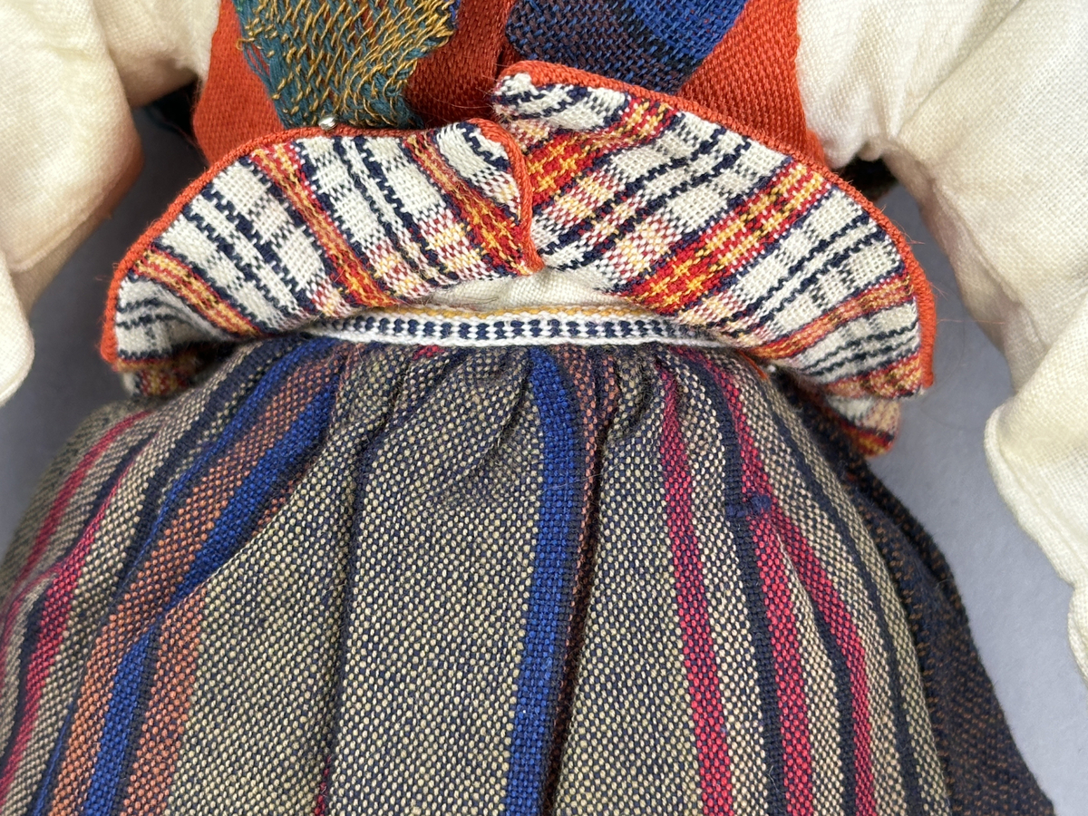 Docka klädd i kvinnlig sockendräkt föreställande Ekshärads socken i Värmland. Huvud och händer sydda av sämskskinn samt kropp av trikåtyg.
