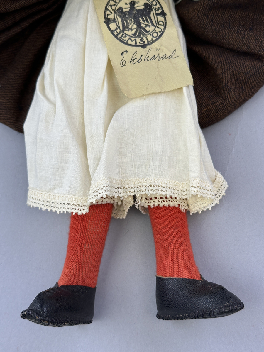 Docka klädd i kvinnlig sockendräkt föreställande Ekshärads socken i Värmland. Huvud och händer sydda av sämskskinn samt kropp av trikåtyg.
