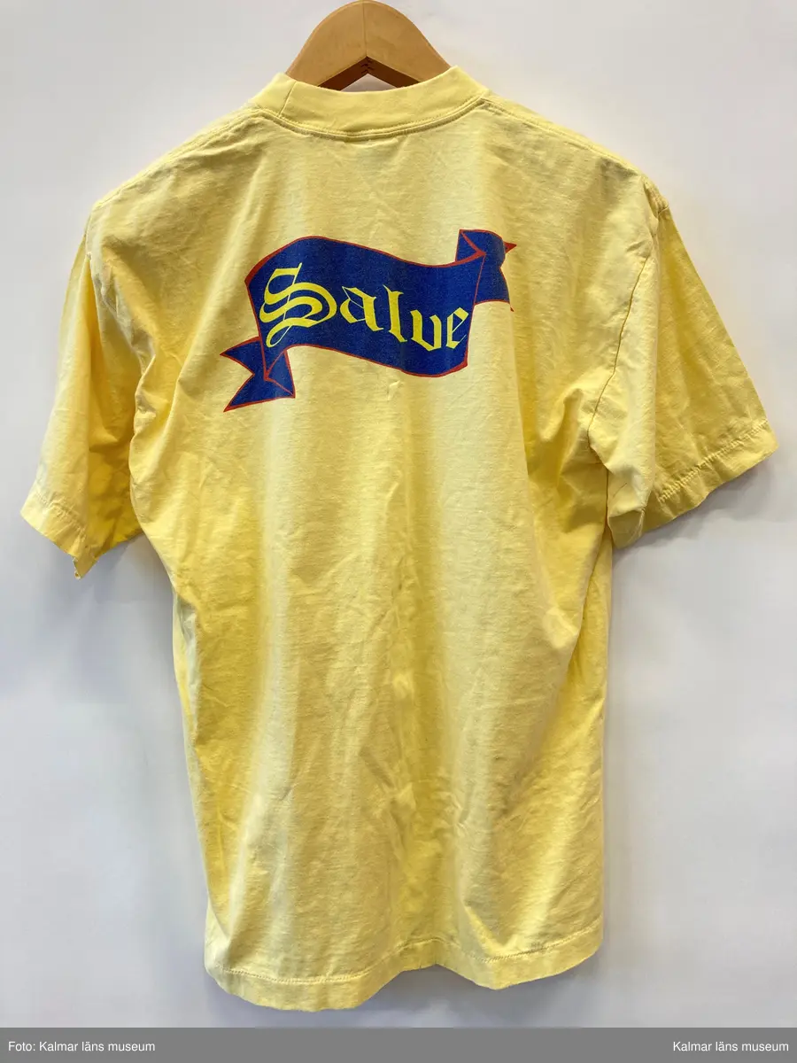 KLM 46894:3. T-shirt. Av bomull i gult med tryckt på ryggen i blått, rött och gult med text: Salve. Storlek large.