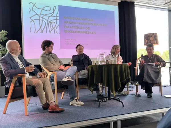 Fem mennesker sitter i lenestoler foran et lite bord på en scene. 