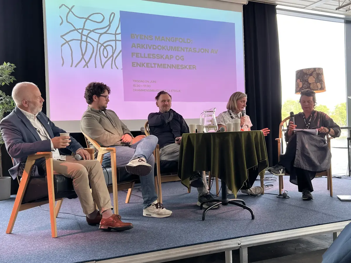 Fem mennesker sitter i lenestoler foran et lite bord på en scene. 