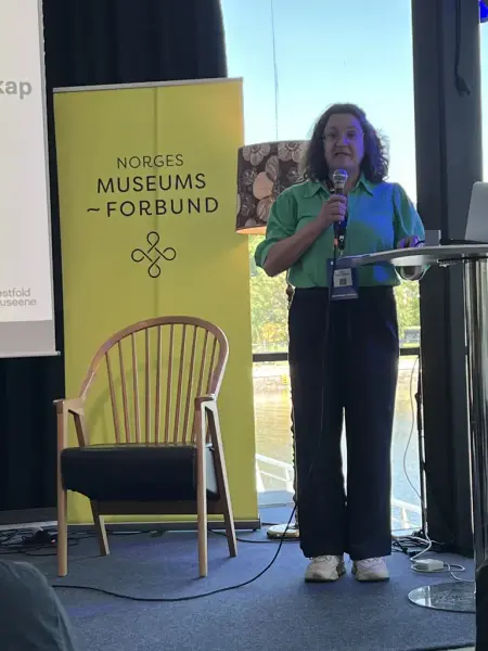 Bildet viser en kvinne i grønn bluse og mørke bukser som står og snakker i en mikrofon foran et høyt bord og en gul roll up med Norges museusmforbund på.