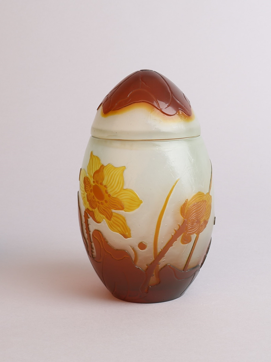 Vase med lokk i melkehvitt opakt overfangsglass. Eggeformet korpus som er dekorert med en gul blomst samt brunlig bladverk i overfangsteknikk. Deler av overflaten er trolig etset.