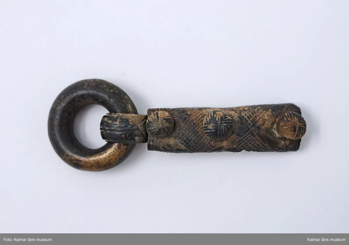 KLM 11783. Bältering med beslag. Sluten ring av grov bronsten. Beslaget närmast cylindriskt försett med tre nitar, ornerade med rutmönster. Beslaget i övrigt har vertikala rutmönster, omslaget om ringen är prytt med linjer, beslaget skadat i ena sidan. En nit lös.