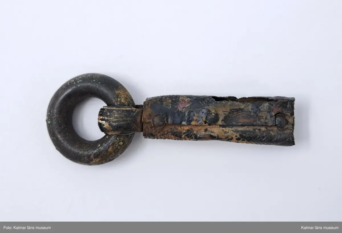 KLM 11783. Bältering med beslag. Sluten ring av grov bronsten. Beslaget närmast cylindriskt försett med tre nitar, ornerade med rutmönster. Beslaget i övrigt har vertikala rutmönster, omslaget om ringen är prytt med linjer, beslaget skadat i ena sidan. En nit lös.