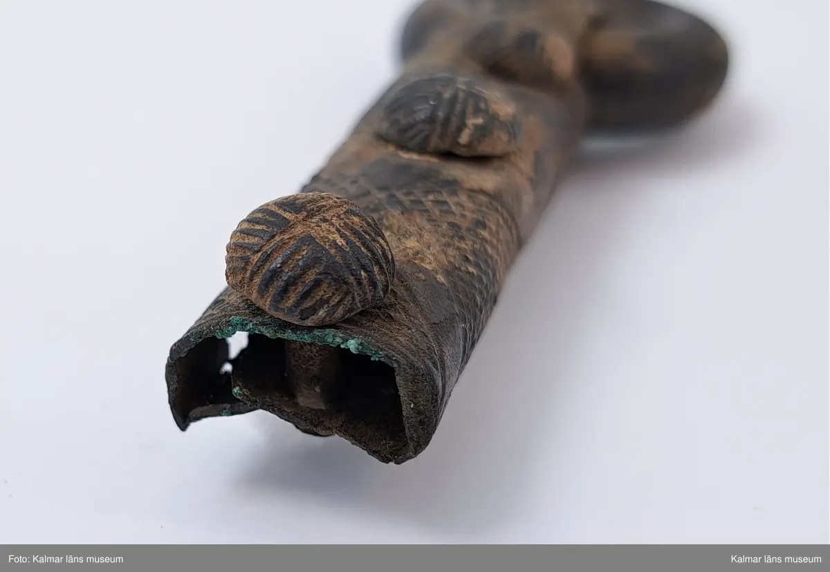 KLM 11783. Bältering med beslag. Sluten ring av grov bronsten. Beslaget närmast cylindriskt försett med tre nitar, ornerade med rutmönster. Beslaget i övrigt har vertikala rutmönster, omslaget om ringen är prytt med linjer, beslaget skadat i ena sidan. En nit lös.
