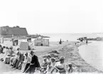 Mycket folk har samlats för att sitta och prata längs strandpromenaden.
I bakgrunden ser man stranden med några badande. Till vänster skymtar Varbergs fästning. 
Något otydligt och suddigt foto.