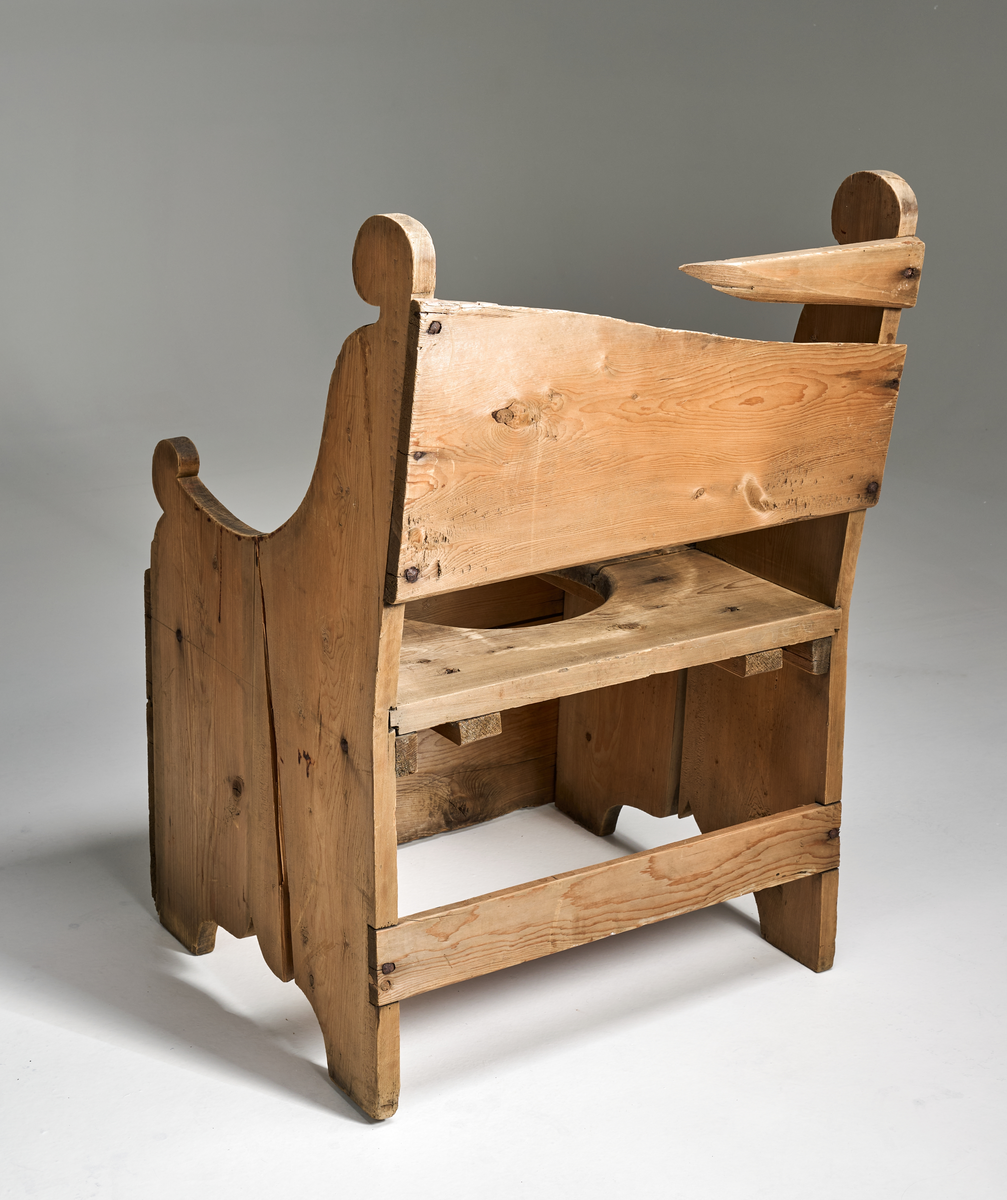 En dostol av ubehandlet tre. Formen er firkantet og den er noe grovt snekret. Setet består av to brede treplanker som er satt sammen ved hjelp av not og fjær. I midten er det skåret ut et sirkulært hull med diameter 29cm. Ryggen består av en bred treplate som er noe ødelagt. Platen dekker ikke hele ryggen. Sidene består av to treplater med enkel utskjæring oppe og nede. I front nede er det festet to brede treplater. Bak nede er det festet en smal treplanke.