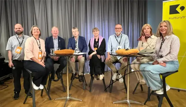 Bildet viser en mann som står og syv personer som sitter på høye stoler. Bak dem står en gul roll up med ordet Kulturytring på