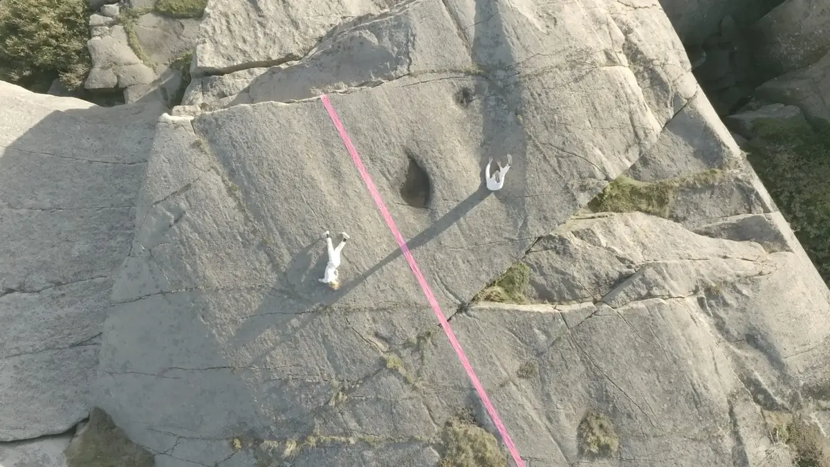 Viser to hvitkledde personer fremføre koreografi, mens de henger i en fjellvegg.