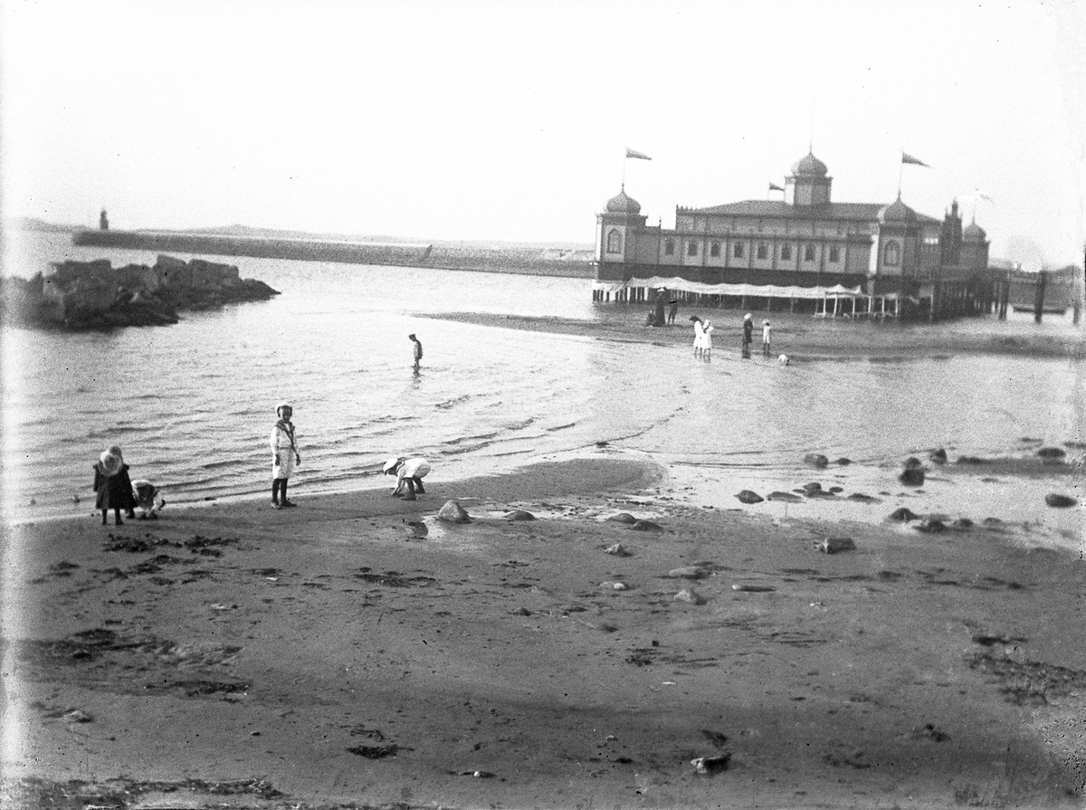 Vy över stranden och kallbadhuset i Varberg. I förgrunden är några barn sysselsatta med att leta snäckor i vattenbrynet.  
Det verkar vara ebb.