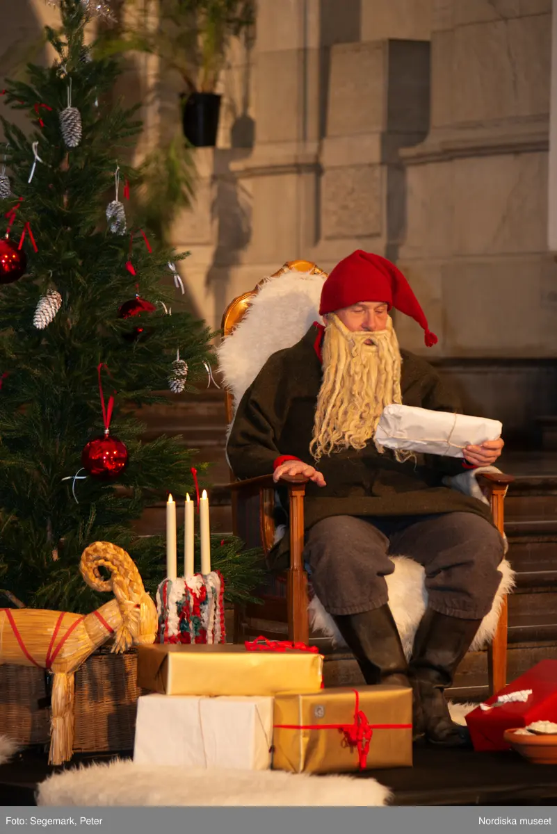 En tomte är på besök i Nordiska museet. Han sitter vid den klädda julgranen och läser på julklapparna. Nedanför granen inslagna paket och en julbock.
