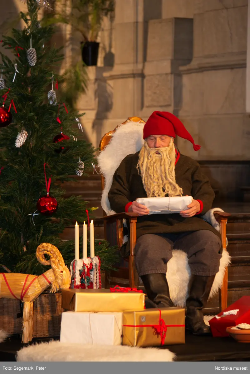 En tomte är på besök i Nordiska museet. Han sitter vid den klädda julgranen och läser på julklapparna. Nedanför granen inslagna paket och en julbock.