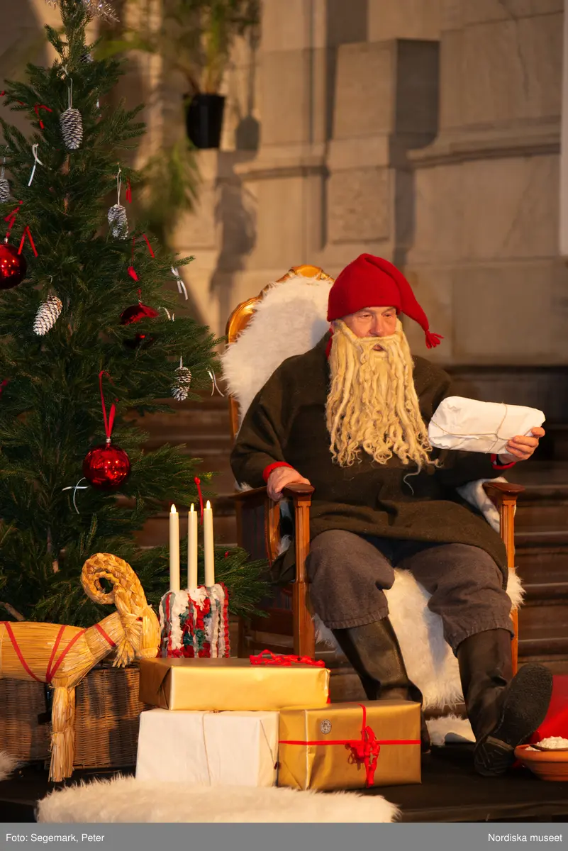 En tomte är på besök i Nordiska museet. Han sitter vid den klädda julgranen och läser på julklapparna. Nedanför granen inslagna paket och en julbock.