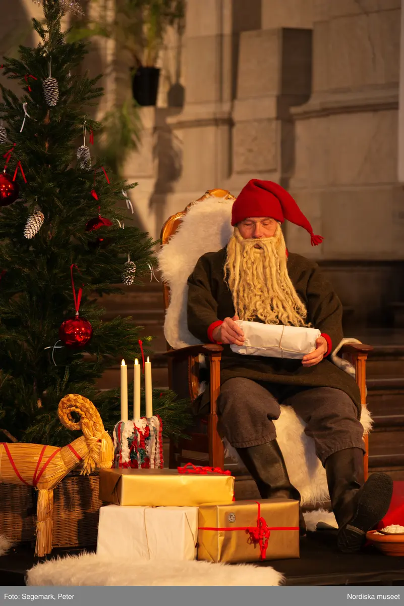 En tomte är på besök i Nordiska museet. Han sitter vid den klädda julgranen och läser på julklapparna. Nedanför granen inslagna paket och en julbock.