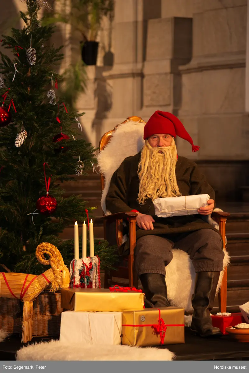 En tomte är på besök i Nordiska museet. Han sitter vid den klädda julgranen och läser på julklapparna. Nedanför granen inslagna paket och en julbock.