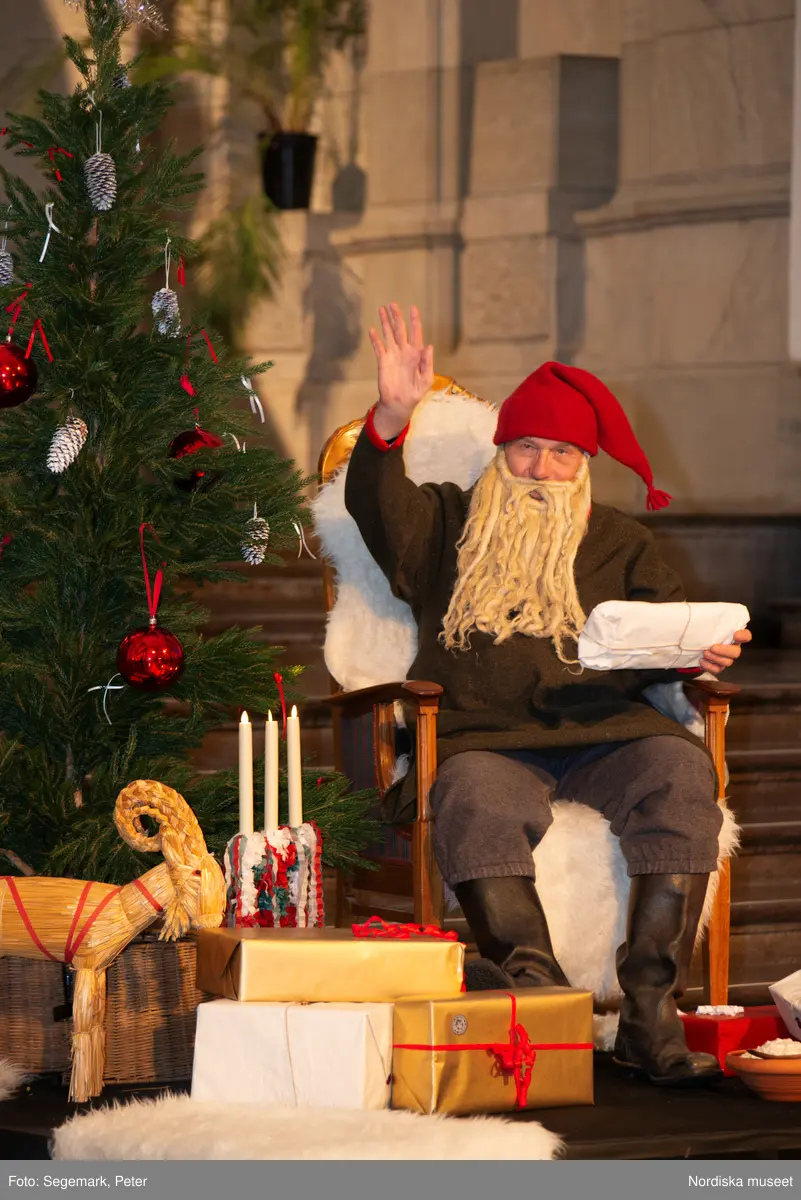 En tomte är på besök i Nordiska museet. Han sitter vid den klädda julgranen och läser på julklapparna. Nedanför granen inslagna paket och en julbock.