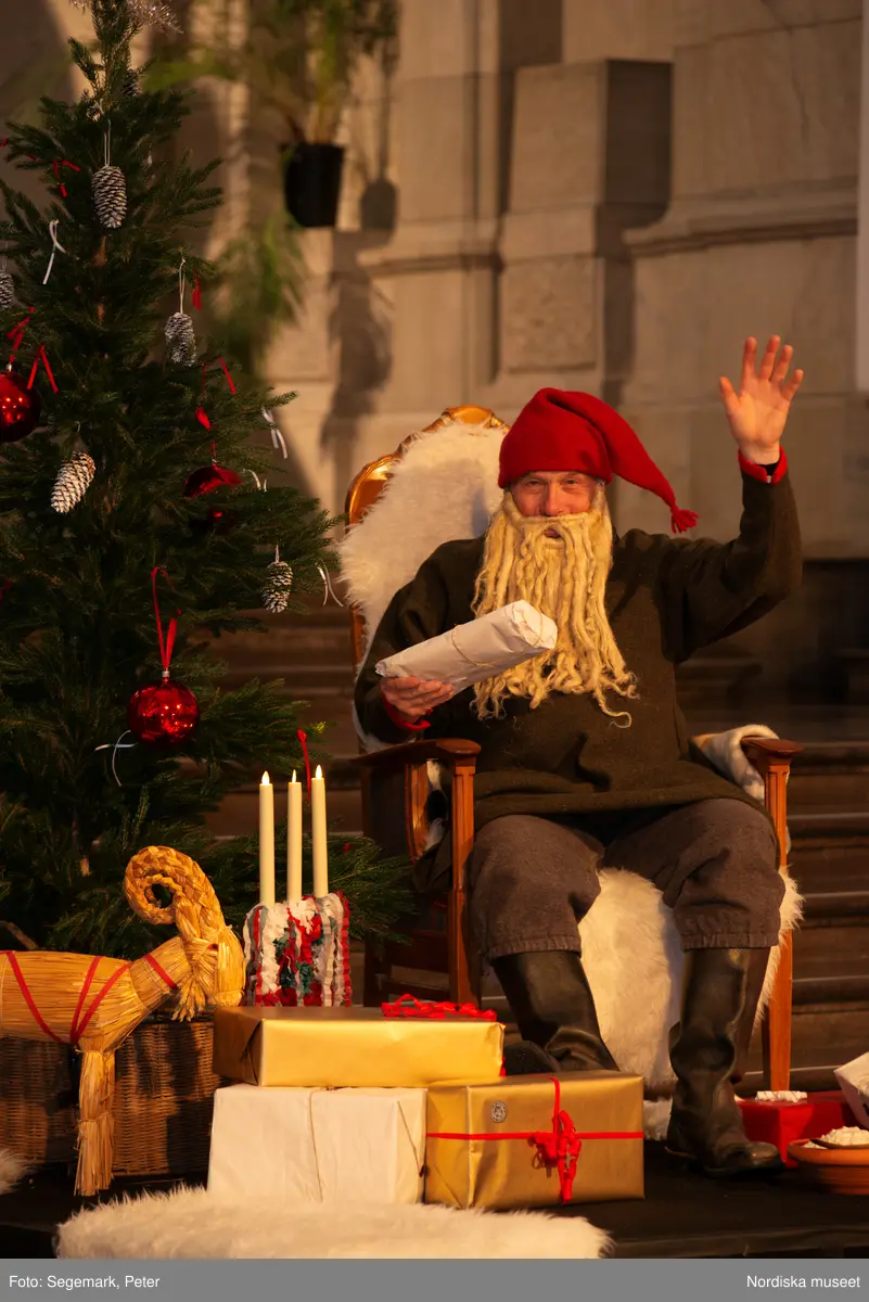 En tomte är på besök i Nordiska museet. Han sitter vid den klädda julgranen och läser på julklapparna. Nedanför granen inslagna paket och en julbock.