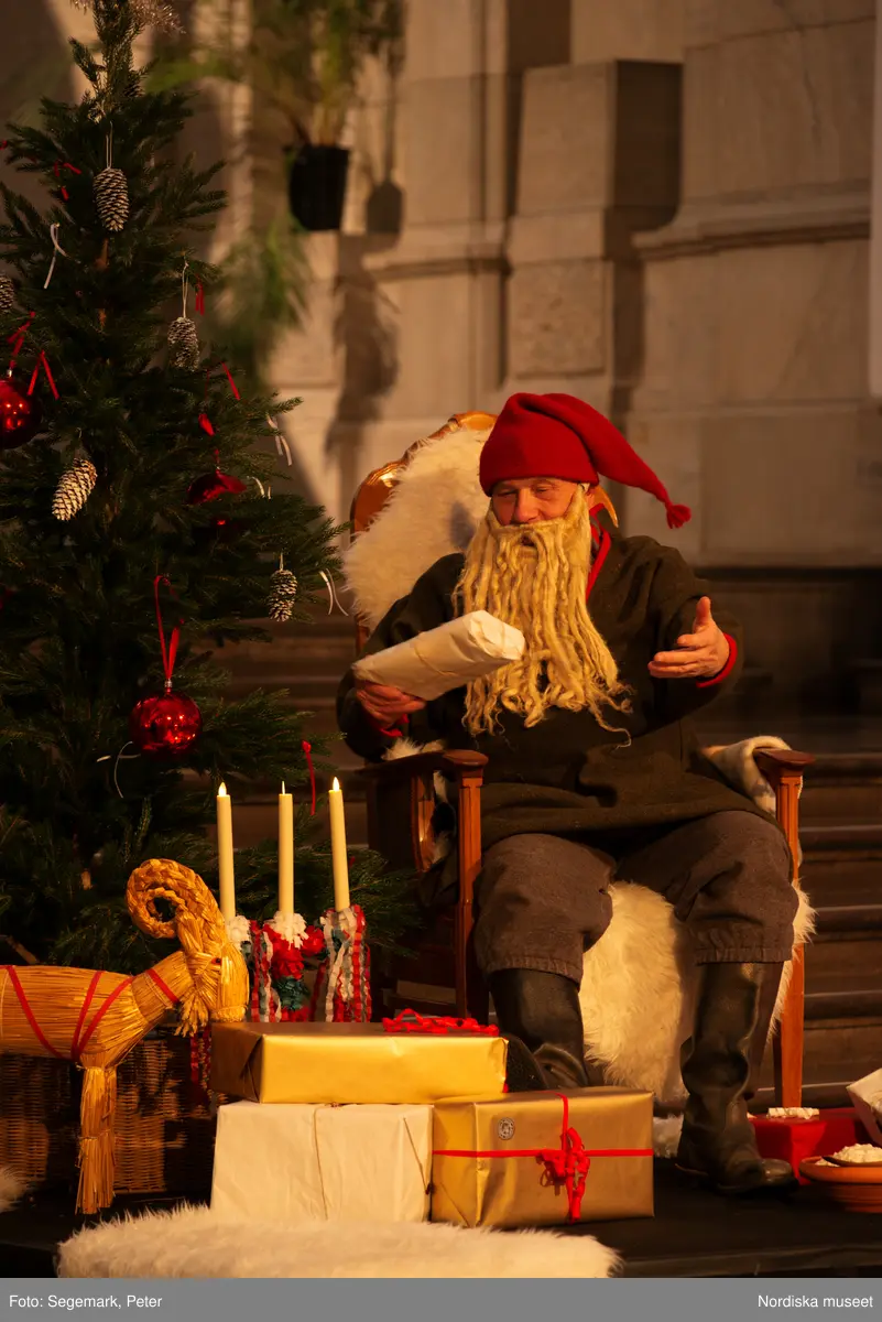 En tomte är på besök i Nordiska museet. Han sitter vid den klädda julgranen och läser på julklapparna. Nedanför granen inslagna paket och en julbock.