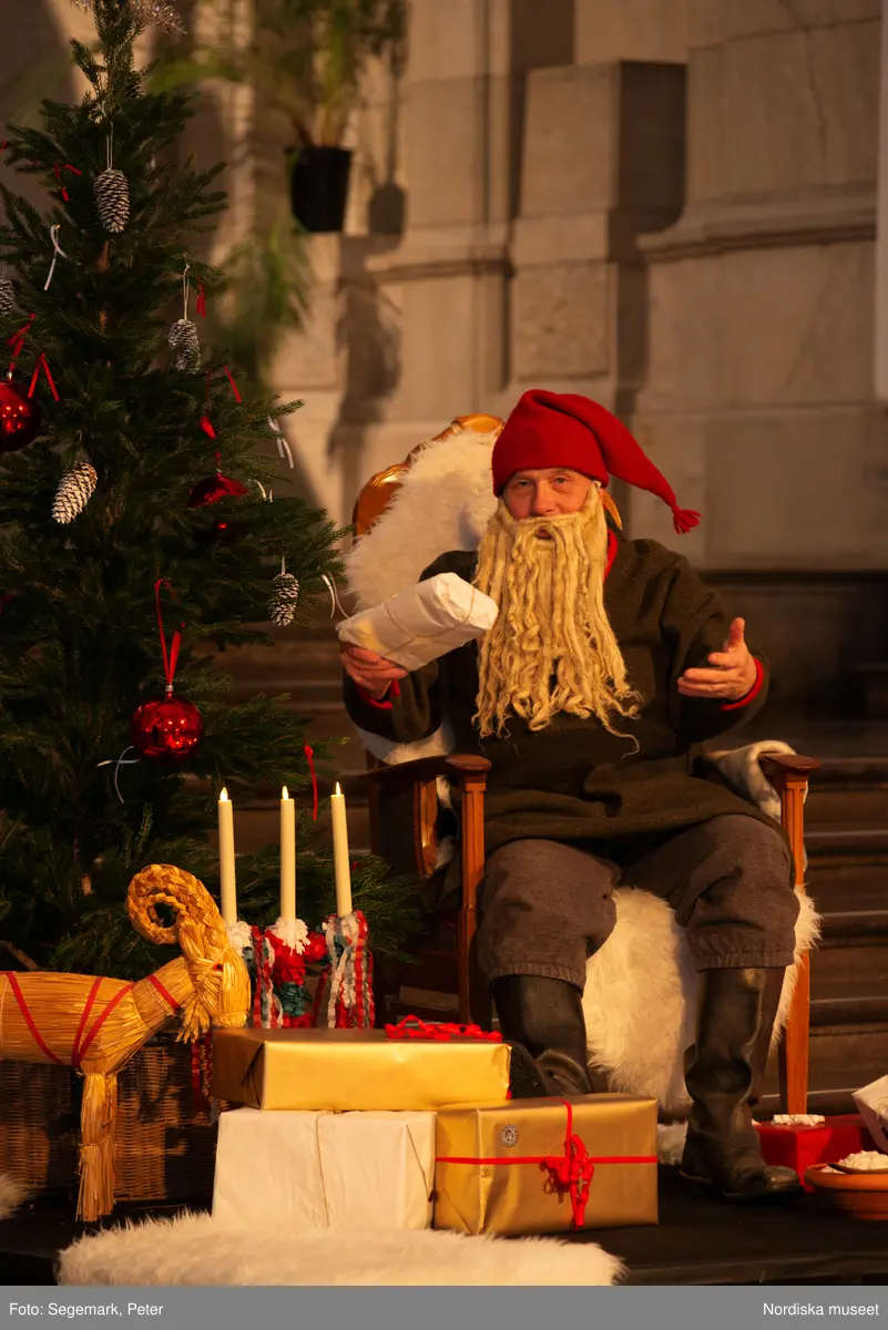 En tomte är på besök i Nordiska museet. Han sitter vid den klädda julgranen och läser på julklapparna. Nedanför granen inslagna paket och en julbock.