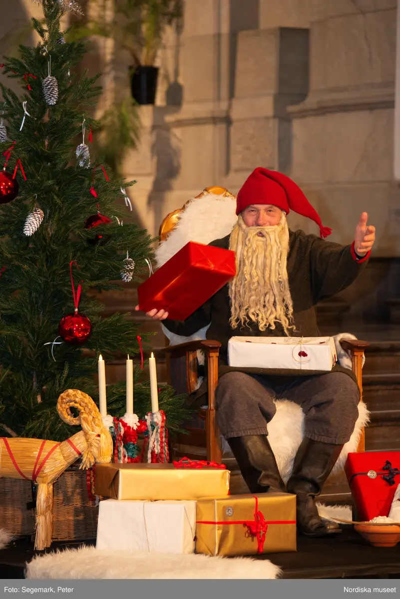 En tomte är på besök i Nordiska museet. Han sitter vid den klädda julgranen och läser på julklapparna. Nedanför granen inslagna paket och en julbock.